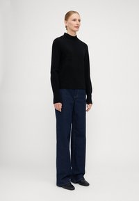 Maglione a collo alto in maglia nera con maniche lunghe, abbinato a jeans a gamba larga blu scuro e scarpe nere senza lacci. Tessuto morbido, design minimal.
