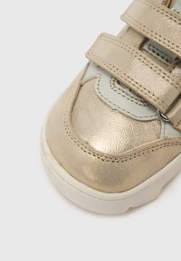 Μεταλλικό χρυσό αθλητικό παπούτσι με λείο, υφασμάτινο πάνω μέρος, διακοσμημένο με ανοιχτό μπλε λεπτομέρειες και διπλούς ιμάντες με κούμπωμα επάνω. Στρογγυλή μύτη και λευκή σόλα.