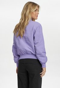 Lavandas krāsas bomber jakas ar pieguļošu ribotu apakšmalu un piedurknēm. Iespējama gluda, nedaudz spīdīga tekstūra un brīva silueta.