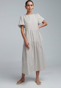 Robe midi sans manches en beige clair avec des rayures foncées, dotée de manches courtes bouffantes et d'une jupe à volants. Portée avec des sandales plates.