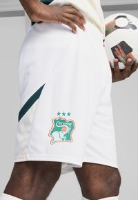 Shorts de football blancs avec le logo brodé de la Côte d'Ivoire en turquoise et orange, présentant un tissu texturé et des accents latéraux noirs. Un ballon de football est tenu.