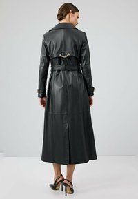 Zwarte leren trenchcoat met een ceintuur in de taille, kraag en kettingdetails aan de achterkant. Lange lengte en gladde textuur, gecombineerd met zwarte hoge hakken.