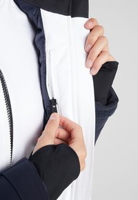 Černobílá zateplená bunda s kapsou na zip na hrudi. Detailní pohled na ruku držící zip a textury látky.