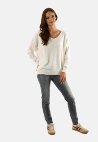 Suéter blanco de cuello en V con mangas largas, combinado con jeans ajustados grises y zapatillas beige con acentos en marrón.