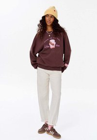 Hnědá mikina s růžovým grafickým textem a potiskem kresleného psa, smetanové kalhoty a hnědé tenisky. Žlutá pletená čepice dokončuje outfit.