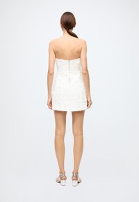 White strapless dress with textured floral pattern, fitted bodice, and short hem. Back zipper detail, paired with silver heeled sandals.