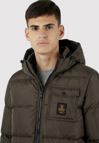 Giacca imbottita verde oliva con chiusura a zip, cappuccio e una tasca sul petto con toppa logo. Tessuto liscio con sezioni trapuntate.
