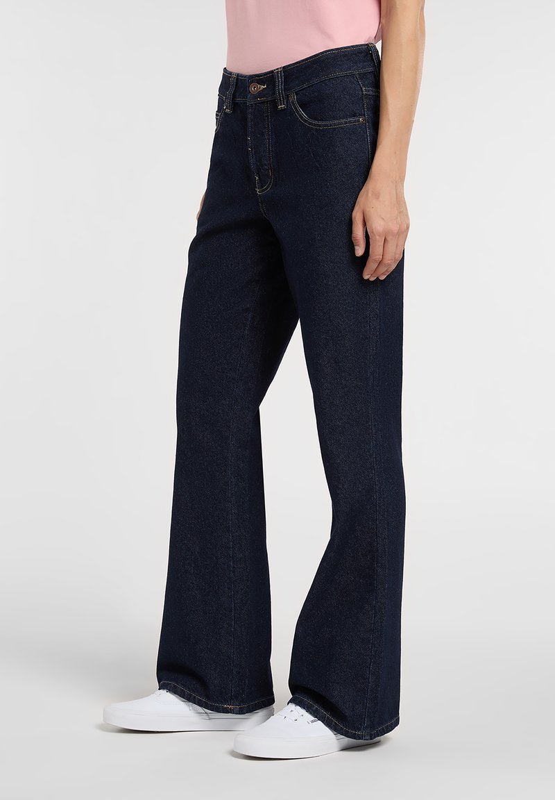 Dark blue denim flared jeans with a mid-rise waist, five-pocket design, and a button fly, paired with white sneakers.