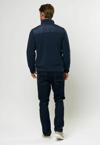 Giacca in pile navy con una parte superiore blu lucido, colletto alto e maniche lunghe, indossata con jeans scuri e scarpe chiare.