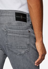 Jeans de denim gris con una superficie texturizada, que presentan una etiqueta negra de Mavi en la cinturilla y un detalle en el bolsillo trasero.