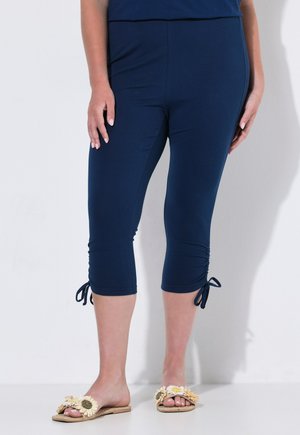 Vrouw draagt marineblauwe cropped leggings met zijstrikken en bloemige slip-on sandalen, staand tegen een witte achtergrond.