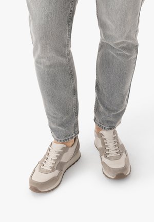 Osoba ubrana w jasnoszare dżinsy i beżowe trampki zamszowymi wstawkami, stojąca na białym tle.