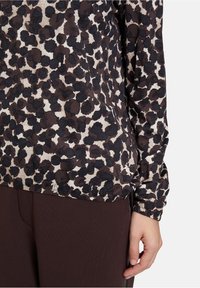 Košile s dlouhým rukávem s leopardím vzorem v hnědé, krémové a černé barvě. Tkanina má texturovaný vzhled, doplněná tmavě hnědými kalhotami.