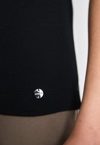 Zwarte ribgebreide top met een zilveren logo label aan de zoom. De stof heeft een gladde textuur en zit strak tegen de huid.