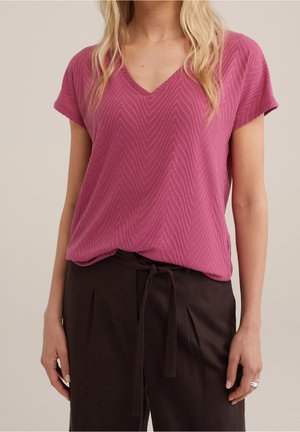 Femme portant un haut rose texturé à col en V et manches courtes, rentré dans un pantalon noir taille haute avec une ceinture nouée à la taille.