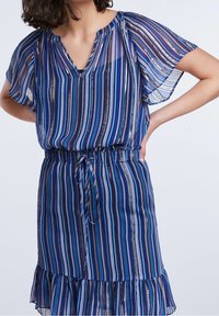 Blusa a righe blu e viola in tessuto trasparente, con maniche corte, scollatura a V e vita cinchita con orlo arricciato.