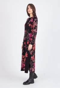 Long black dress with floral pattern in shades of red and pink, featuring long sleeves and side slits. Worn with black ankle boots.