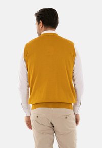 Gilet lavorato a maglia giallo senape con scollo alto, indossato sopra una camicia bianca a maniche lunghe. Presenta un design aderente e un orlo inferiore a coste.