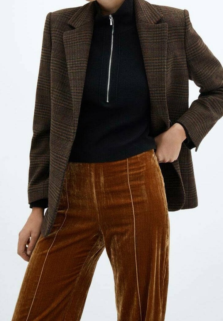Personne portant un blazer à carreaux marron sur un haut noir zippé et un pantalon en velours marron taille haute avec des coutures verticales visibles.