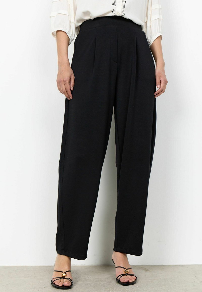 Femme portant un pantalon noir taille haute à jambes larges, associé à des sandales noires à brides et un chemisier blanc à manches 3/4.
