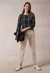 Schwarz-weiße karierte Langarmbluse, beiges Zugband-Hose und schwarze Daunenjacke in der linken Hand, kombiniert mit weißen Sneakers.