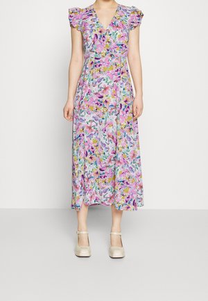 Femme portant une robe fleurie sans manches, à encolure en V, longueur cheville, avec des épaules à volants et des chaussures Mary Jane couleur crème.