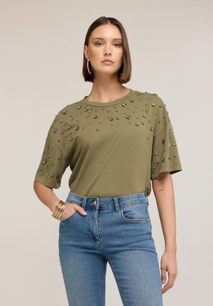 Junge Frau mit kurzen braunen Haaren, trägt ein olivgrünes, mit Nieten besetztes T-Shirt und blaue Jeans, steht mit einer Hand in der Tasche vor einem beigen Hintergrund.