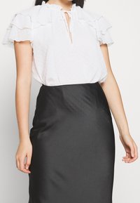 Kvinna i en vit texturerad kortärmad blus med volanger på ärmarna och en svart högmidjad sidenkjol, stående med avslappnade armar.