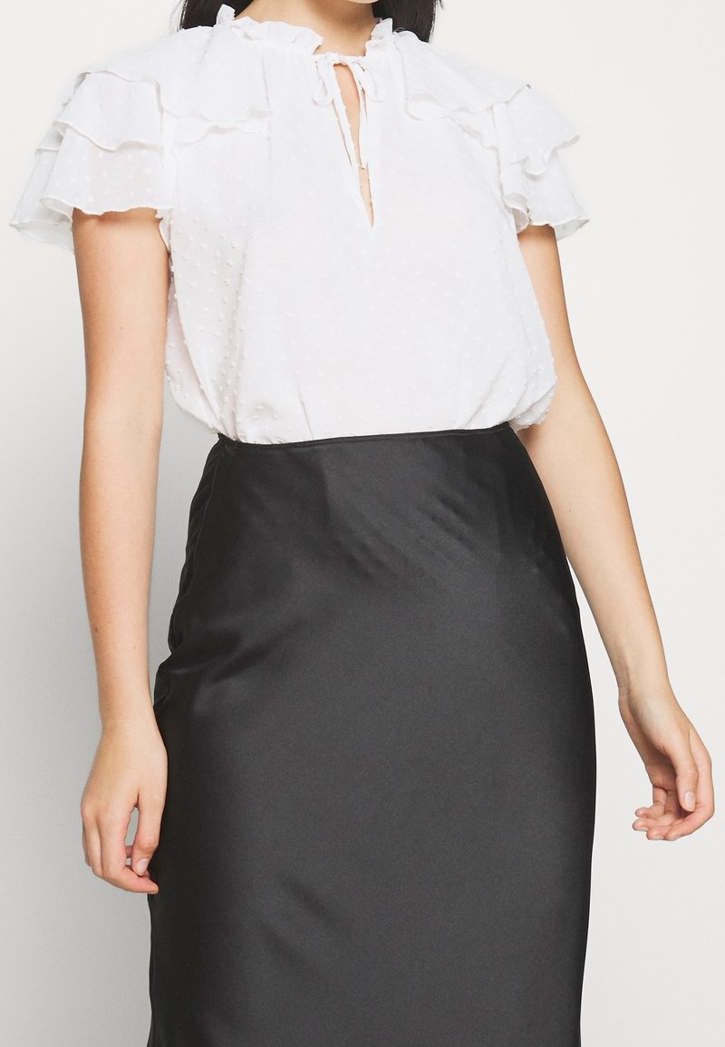 Kvinna i en vit texturerad kortärmad blus med volanger på ärmarna och en svart högmidjad sidenkjol, stående med avslappnade armar.