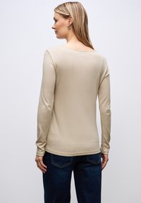 Beige Langarmshirt mit rundem Halsausschnitt, aus weichem Stoff gefertigt. Hat enganliegende Ärmel und einen leicht geschwungenen Saum. Kombiniert mit dunklen Jeans.