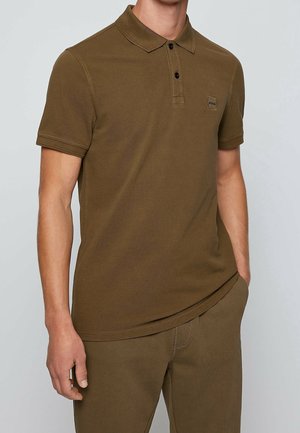 Man draagt een bruin poloshirt met korte mouwen en twee knopen en een bijpassende bruine broek, staat met één hand in de zak.
