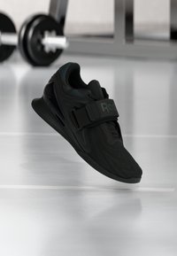 Μαύρο αθλητικό παπούτσι Reebok για άρση βαρών με υφή στην επιφάνεια και λουράκι, αιωρείται πάνω από το πάτωμα γυμναστηρίου με φόντο θολό μπάρα βάρη.