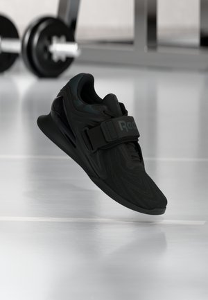 Schwarzer Reebok Gewichtheberschuh mit strukturiertem Obermaterial und Riemen, schwebend über dem Fitnessboden mit unscharfer Langhantel im Hintergrund.