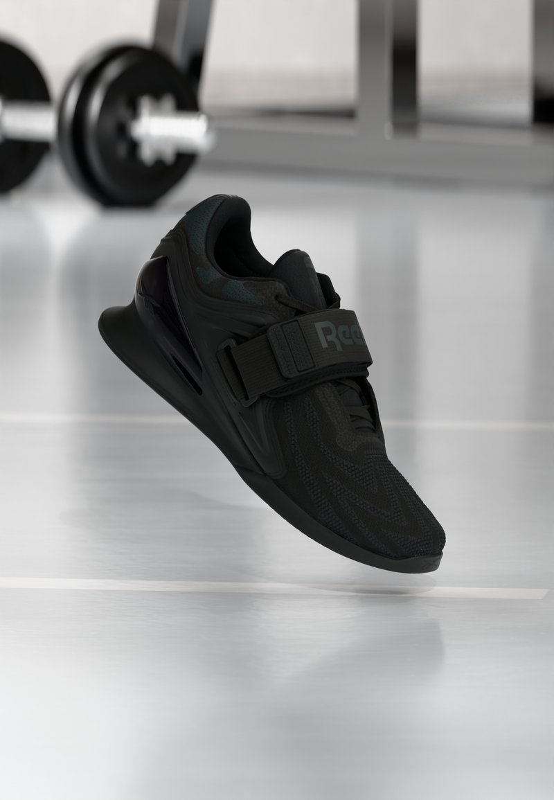 Sort Reebok-vægtløftersko med tekstureret overdel og rem, svævende over gymnastikgulv med sløret vægtstang i baggrunden.