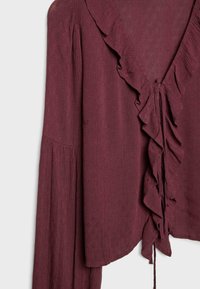 Blouse bordeaux en tissu texturé, avec un décolleté en V profond, des volants, des manches longues et un détail à nouer à la taille.