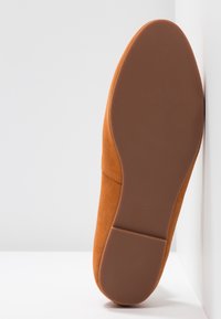 Semelle plate de chaussure marron face au spectateur, reposant verticalement contre un mur d'angle blanc sur une surface blanche.