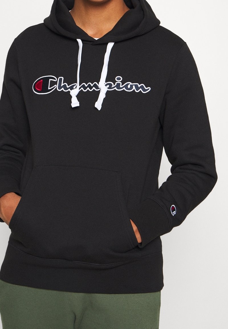 Personne portant un sweat à capuche Champion noir avec des cordons blancs, logo sur la poitrine et la manche, les mains dans la poche avant, assorti d'un pantalon vert.