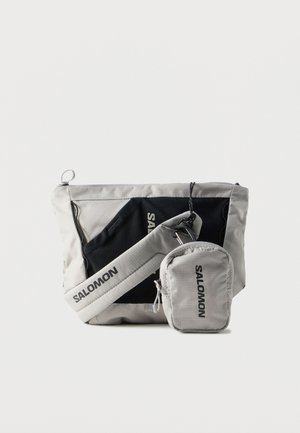 Grijze nylontas met een gestructureerde afwerking, voorzien van een zwart paneel, verstelbare riem en een klein zakje met rits. Voorzien van de "SALOMON" branding.