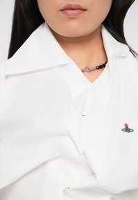 Cămașă albă cu guler, având o textură șifonată, cu un mic design brodat. Colier din mărgele negre cu închidere din argint.