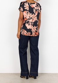 Kvinde iført en sort og ferskenfarvet blomstret kortærmet bluse, mørkeblå vide jeans og sorte hæle-sandaler, stående foran en hvid væg.