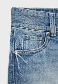 Gros plan d’un jean en denim bleu montrant la ceinture avant, la boucle de ceinture, les rivets et une petite poche à monnaie avec des détails de couture visibles.