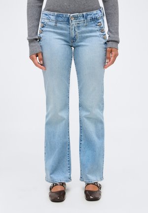 Personne portant un jean flare bleu clair avec des détails de boutons sur le côté, un haut gris à manches longues côtelé, et des chaussures plates marron foncé avec des brides.