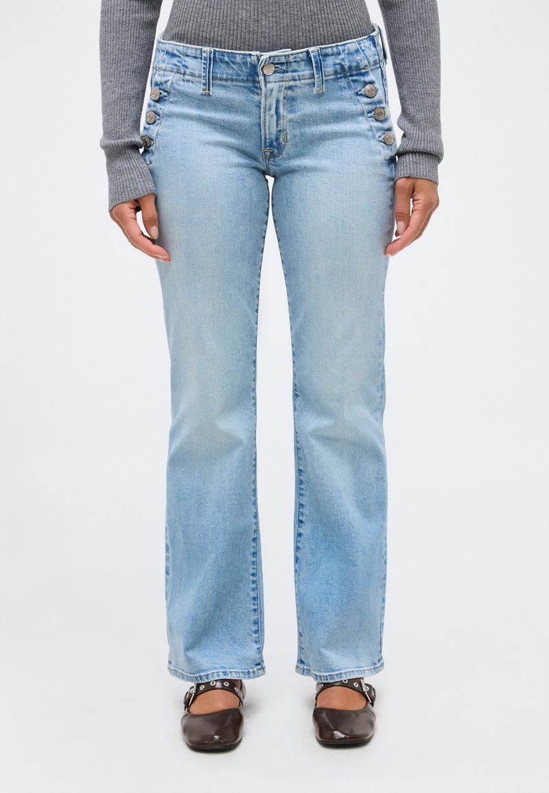 Άτομο που φοράει ανοιχτό μπλε τζιν με απόληξη καμπάνα και λεπτομέρειες με κουμπιά στο πλάι, γκρι ριμπ πλεκτή μπλούζα με μακριά μανίκια και σκουρό καφέ επίπεδα παπούτσια με λουράκια.