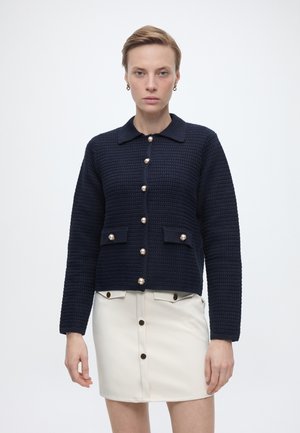 Γυναίκα με κοντά μαλλιά που φοράει μια μπλε πλεκτή ζακέτα με κουμπιά από πέρλες και λευκή φούστα με λεπτομέρειες κουμπιών στο μπροστινό μέρος.