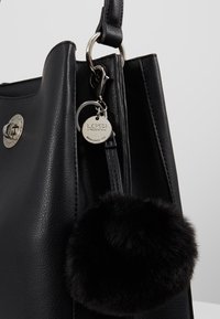 Sac à main en cuir noir avec fermoir argenté, anneau en métal, étiquette de marque ronde et porte-clés pompon noir pelucheux attaché sur fond gris.