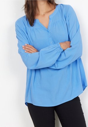 Vrouw draagt een loszittende lichtblauwe blouse met knopen en zwarte broek, armen gekruist voor.