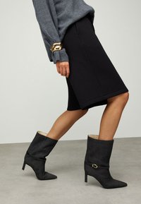 Bottes noires montant jusqu'aux genoux avec des bouts pointus, un talon effilé, une doublure intérieure contrastante ; tissu texturé et une boucle décorative en accent.