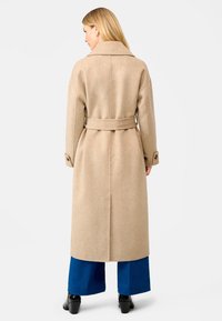 Beige wollen overcoat met een ceintuur in de taille, grote kraag en knoopmouwen, gedragen over wijd blauwe broeken en zwarte laarzen met hak.