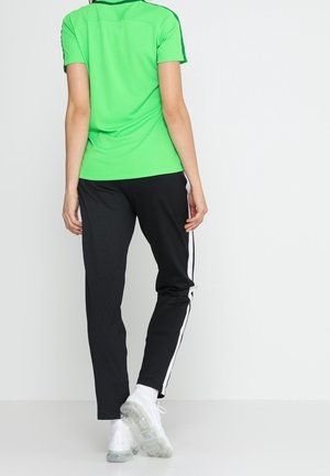 Person trägt ein leuchtend grünes kurzärmeliges Shirt, schwarze Jogginghose mit weißen Seitenstreifen und weiße Turnschuhe, steht mit dem Rücken zur Vorderseite.