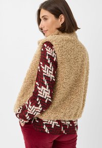 Beige fleecvest met een gestructureerde afwerking, gecombineerd met een multikleurige gestreepte shirt. Het shirt heeft een geometrisch ontwerp in rood, roomwit en zwart.